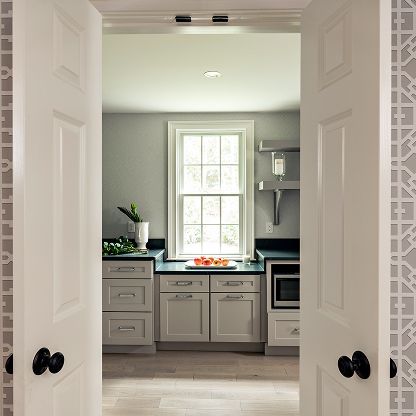 Bright kitchen seen through open double doors, with white cabinets, gray walls, and a window above the sink.