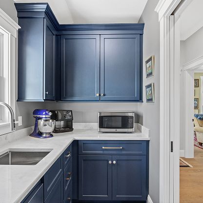 Blue kitchen cabinets with white countertops, sink, stand mixer, and microwave in a bright nook.