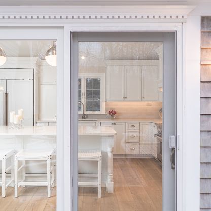 Bright white kitchen viewed through open doorway, with island, cabinets, wood floor, and tiled backsplash.
