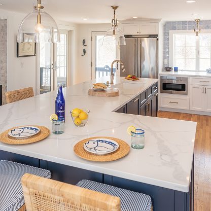 Bright modern kitchen with white island, wicker stools, stainless appliances, and blue table setting.