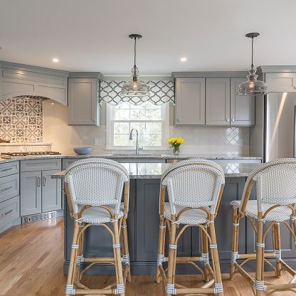 Bright modern kitchen with gray cabinets, white tile, island, and three woven bar stools.