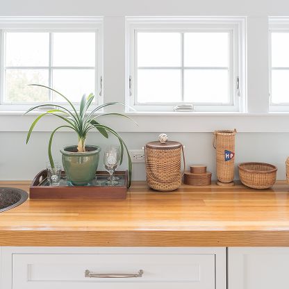 White kitchen countertop with potted plant, wicker baskets, and wooden trays beneath two windows