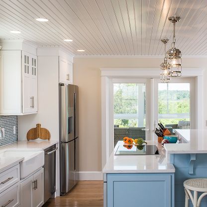 Bright white kitchen with blue island, stainless fridge, pendant lights, and fruit bowl by a window.