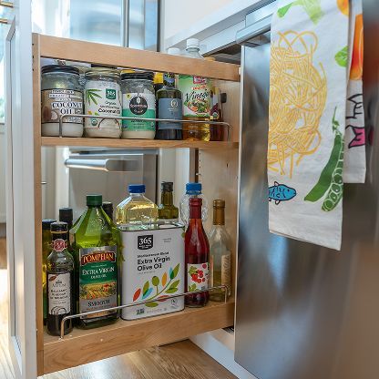 Wooden wall-mounted spice rack with jars and bottles beside a stainless steel refrigerator and tea towel