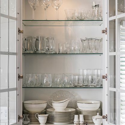 Open kitchen cabinet with glassware and stacked white dishes on glass shelves.