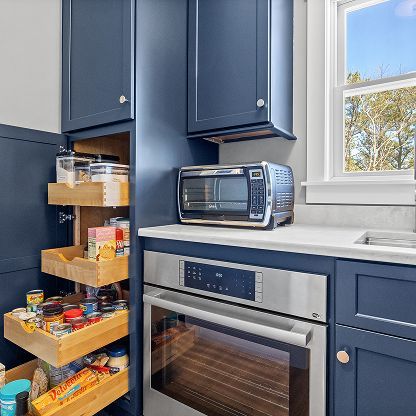Blue kitchen with open pantry drawers, a countertop microwave, and a stainless oven by a window