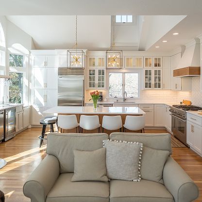 Bright modern kitchen and dining area with white cabinets, island, stools, and a gray sofa in the foreground