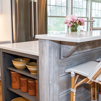 Modern kitchen island with marble countertop, wicker stool, open shelves, and pink flowers by a window