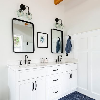 Bright white bathroom vanity with double sinks, black mirrors, wall lights, and white paneled walls