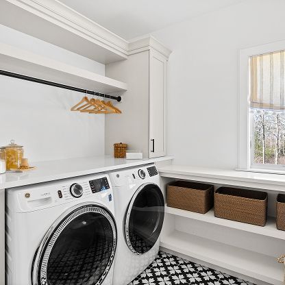 Bright laundry room with front-load washer and dryer, white shelves, and wicker storage baskets by a window.