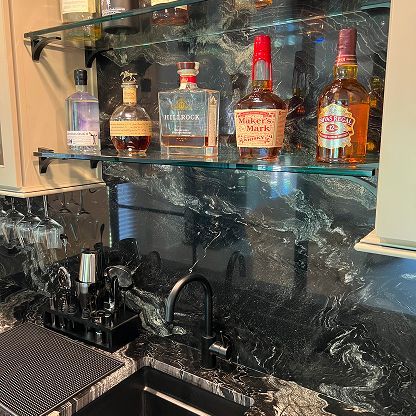 Home bar shelf with liquor bottles above a black sink and marbled backsplash