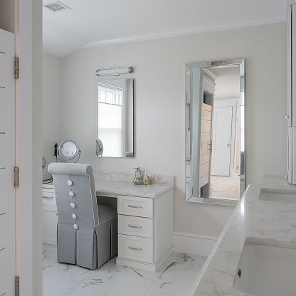 Bright white bathroom with vanity, chair, and large mirrors; doorway reflected in mirror.