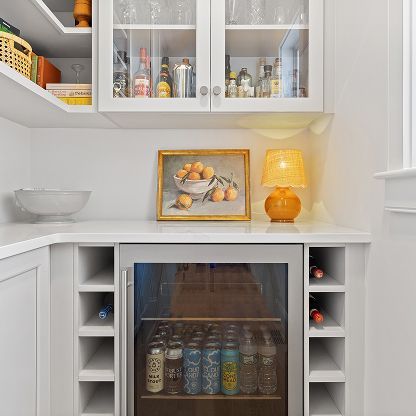 Bright white kitchen nook with glass cabinets, a small drink fridge, fruit painting, and an orange lamp