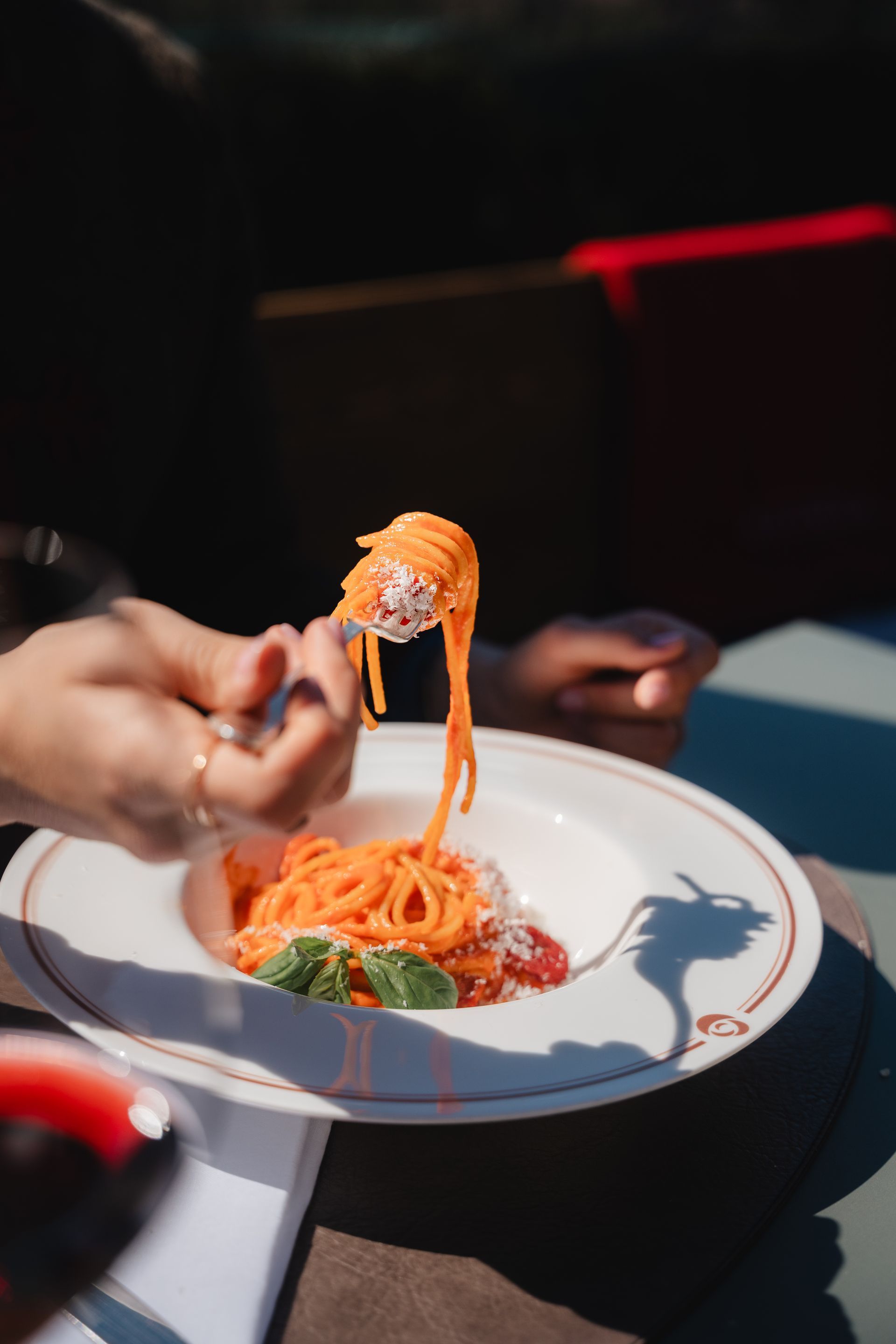 Hand holding a forkful of spaghetti over a white plate at an outdoor table, with sunlight and shadows