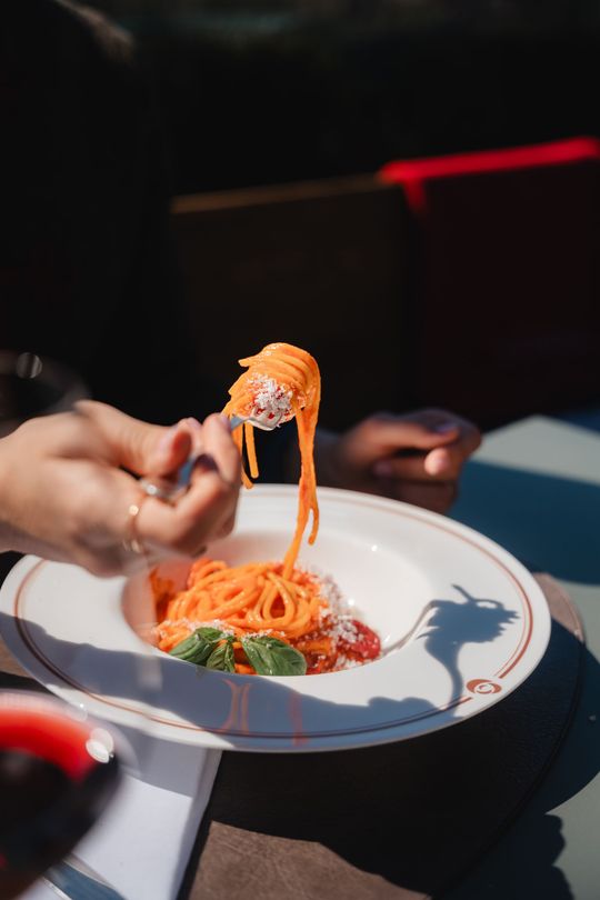 Una mano che stringe una forchettata di spaghetti sopra un piatto bianco in un ristorante scarsamente illuminato.