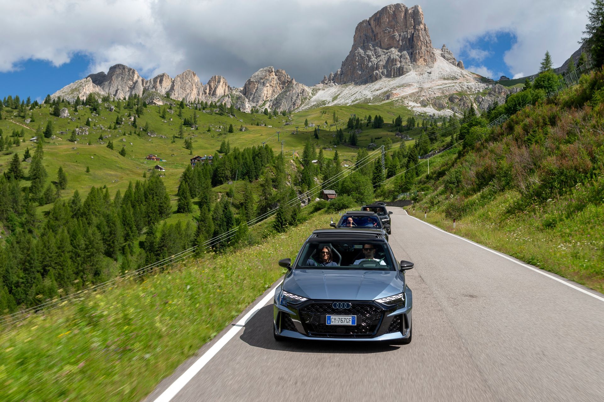 An Audi drives along a mountain road, amidst pine trees and snow-capped peaks, under a clear sky.