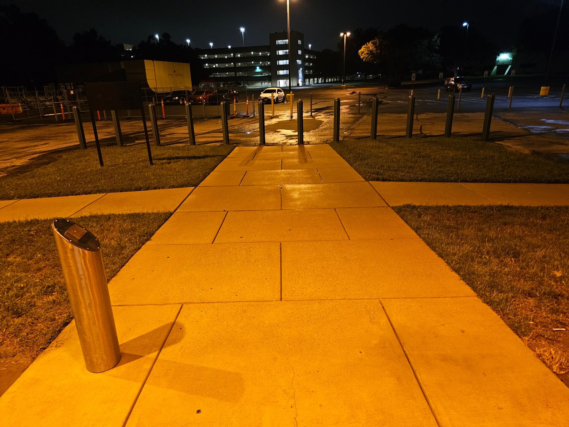 Nighttime walkway lit by streetlights leading to a building, with a short wooden post in the foreground.