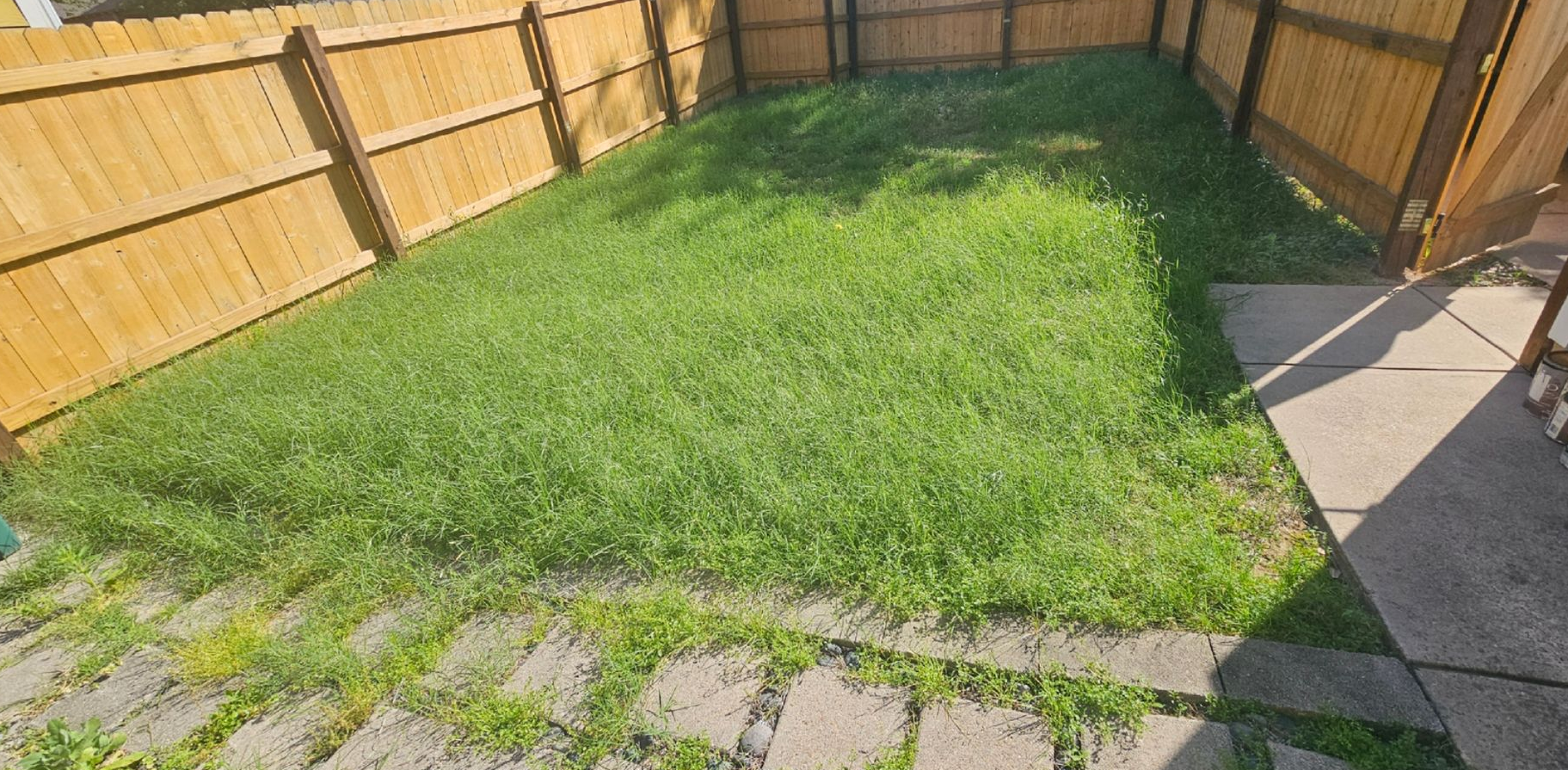 Small fenced backyard with a grassy lawn, stone pavers, and wooden fences on both sides.