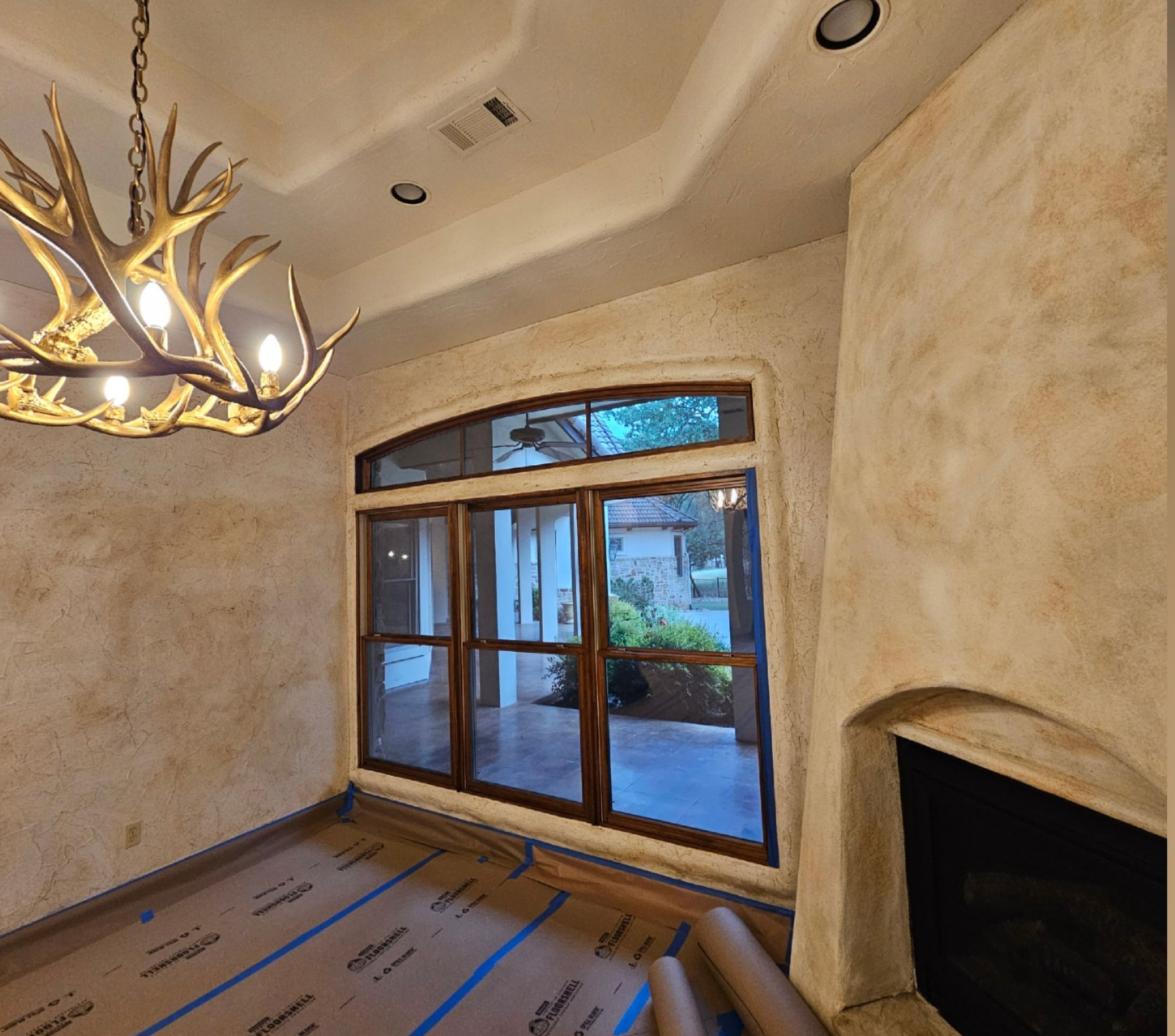 Empty living room with stone fireplace, large window, antler chandelier, and floor covered for renovation