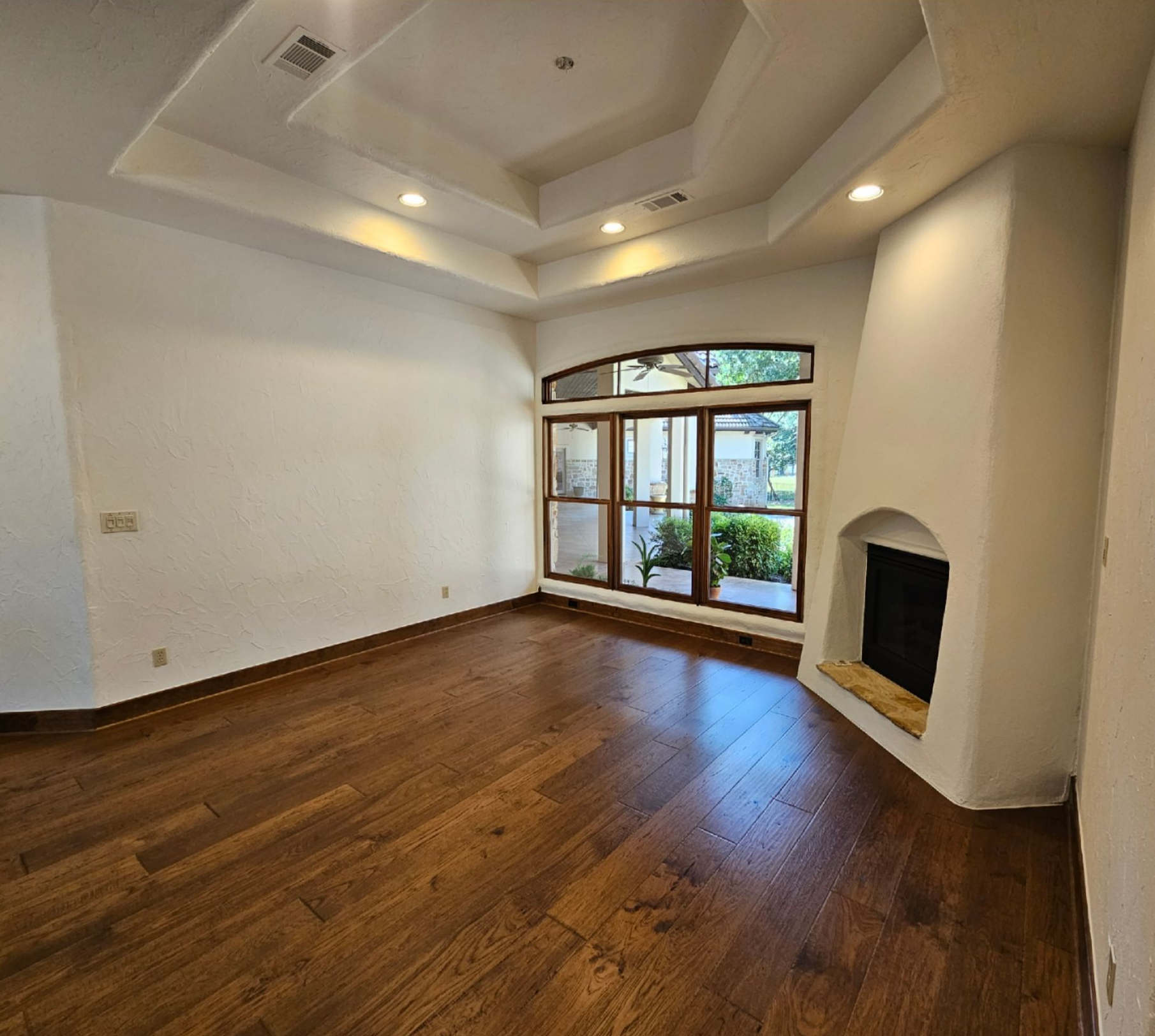Empty living room with hardwood floors, recessed ceiling lights, fireplace, and large windowed doorway.