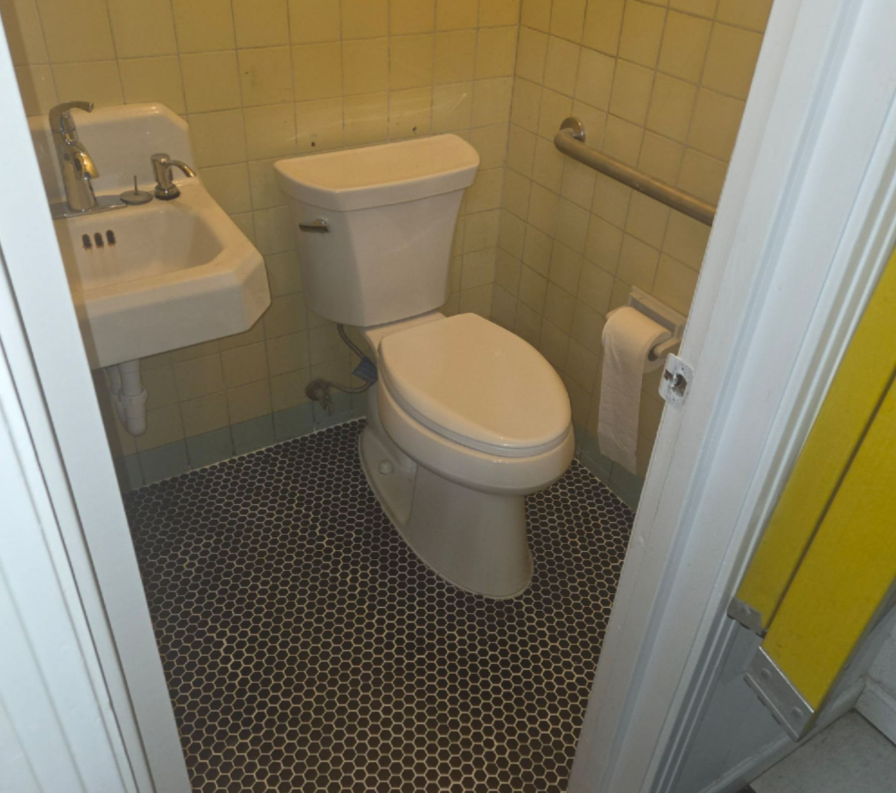 Small bathroom with a toilet, wall-mounted sink, and yellow-tiled walls on a black patterned floor.