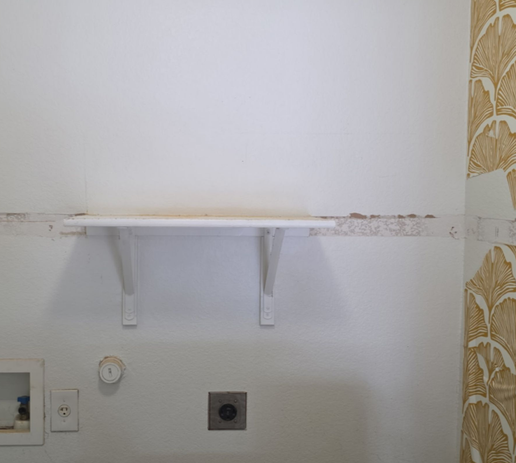 Empty wall with a small white shelf, electrical outlet, and patterned tile on the right; slight wall staining.