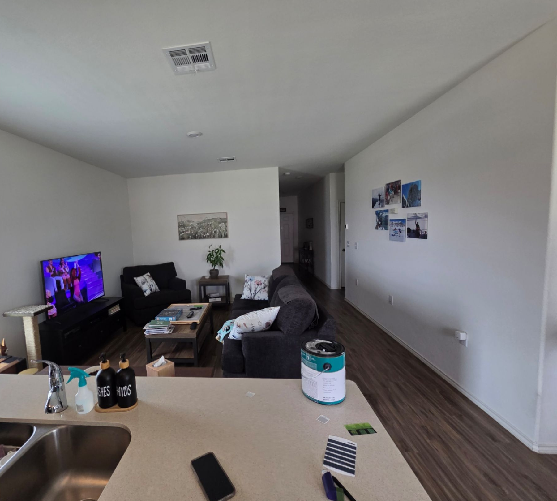 Open-plan living room with a TV, couch, coffee table, wall photos, and a kitchen counter in the foreground