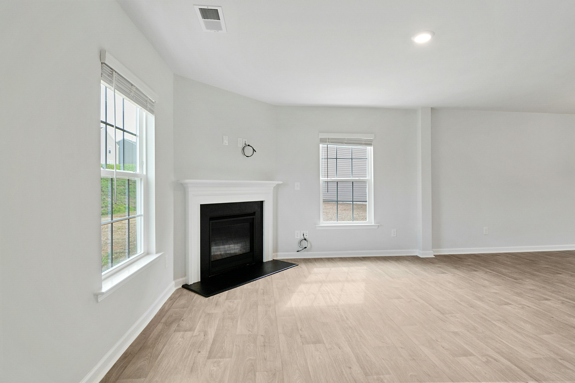 Bright empty living room with light wood floors, white walls, a black fireplace, and two windows.
