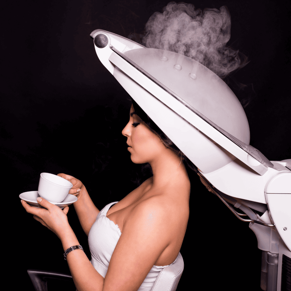 Woman seated under a large steaming hood dryer, holding a white teacup and saucer on a dark background