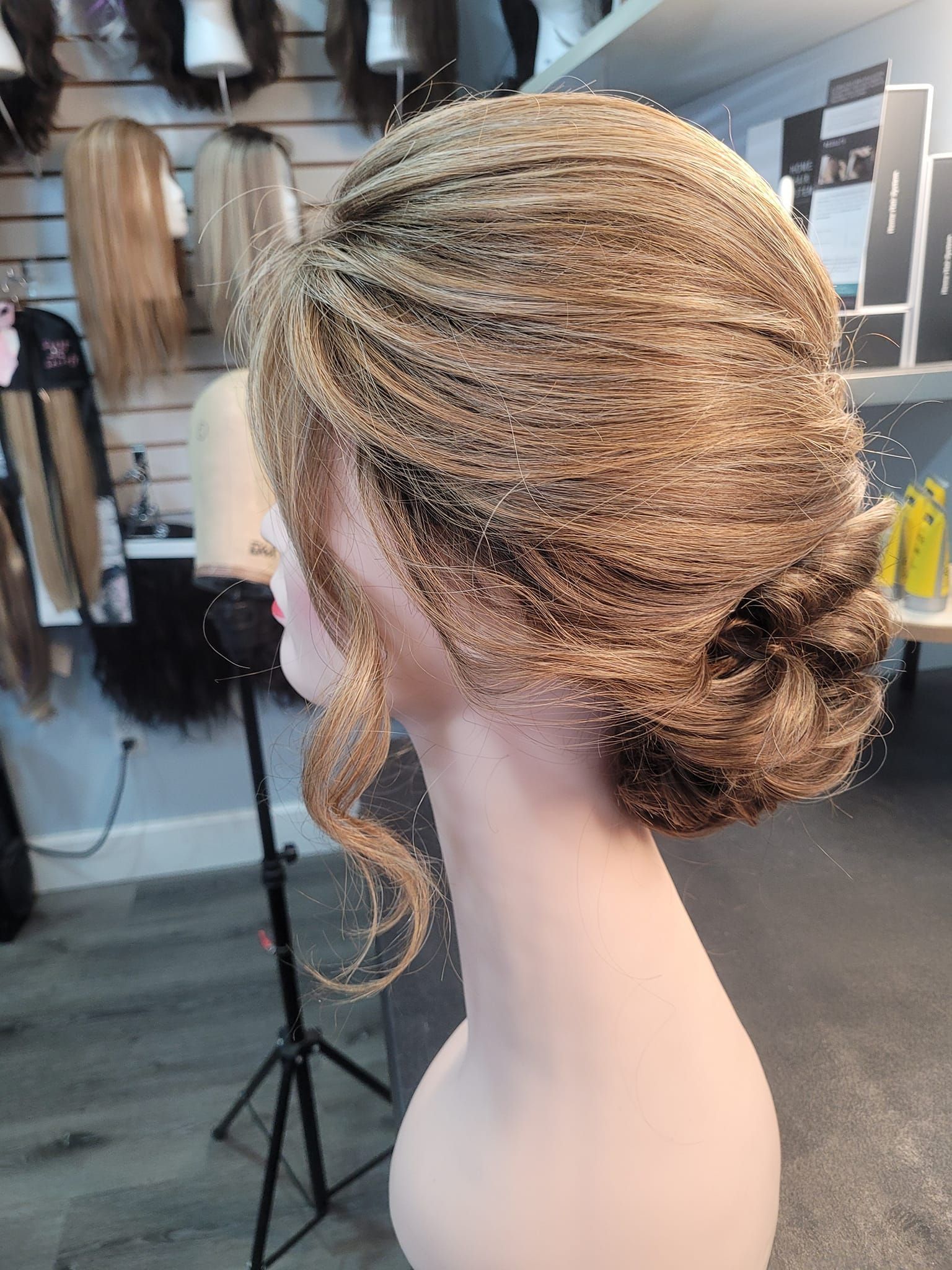 Braided blonde updo wig on a mannequin head in a wig shop, with hanging hairpieces in the background