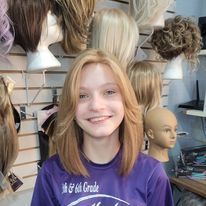 Smiling person with blonde hair in a purple shirt, seated in a wig shop with mannequin heads and wigs behind them