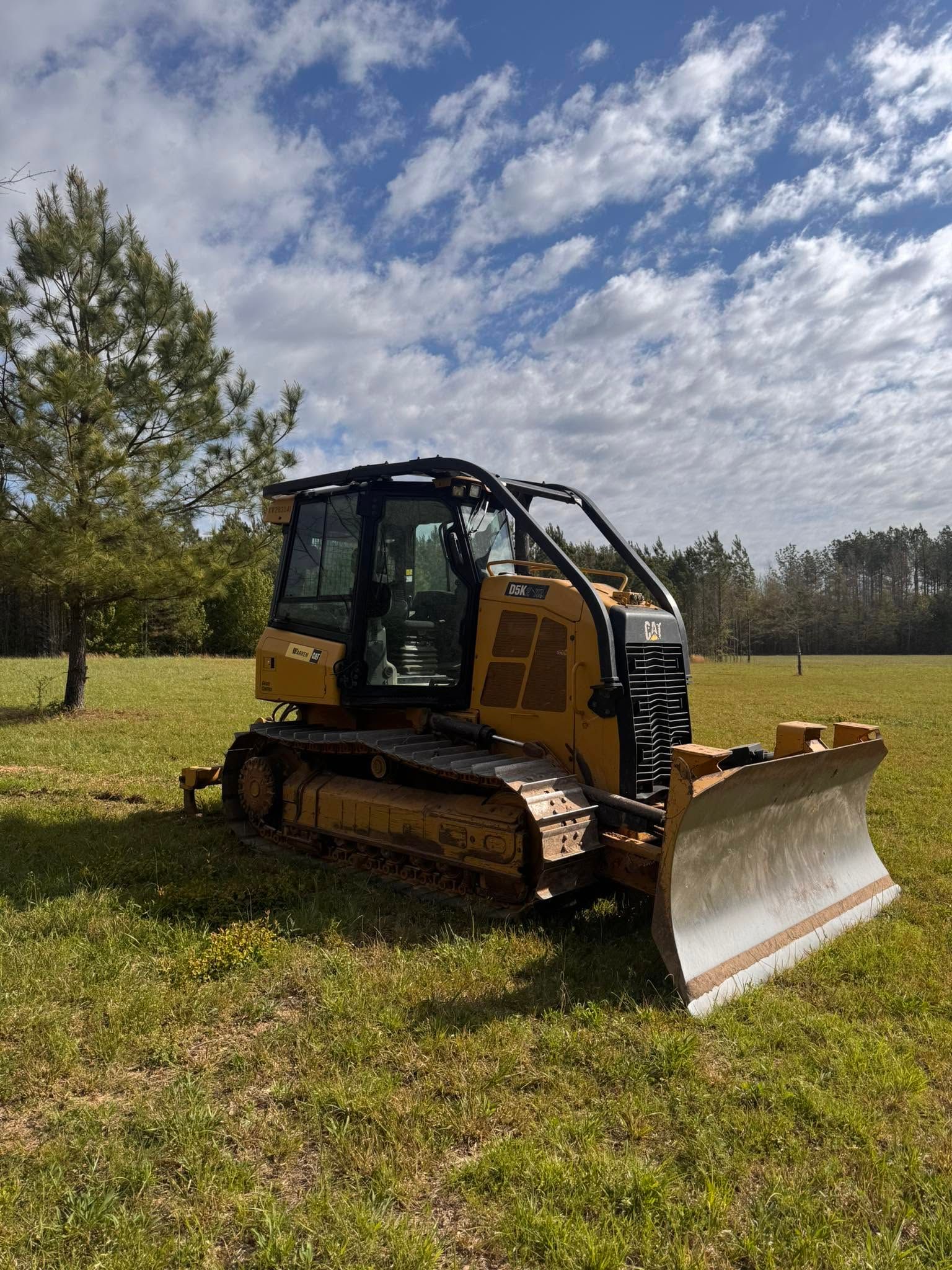 Yellow tracked bulldozer with a front blade parked in a grassy field under a partly cloudy sky