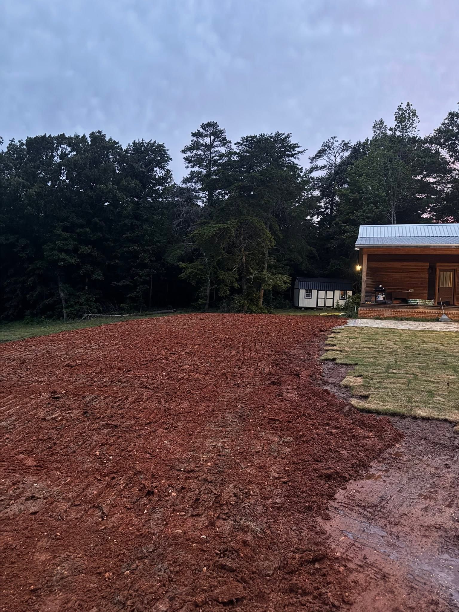 Freshly spread red mulch beside a wooden building at dusk, with trees lining the background.