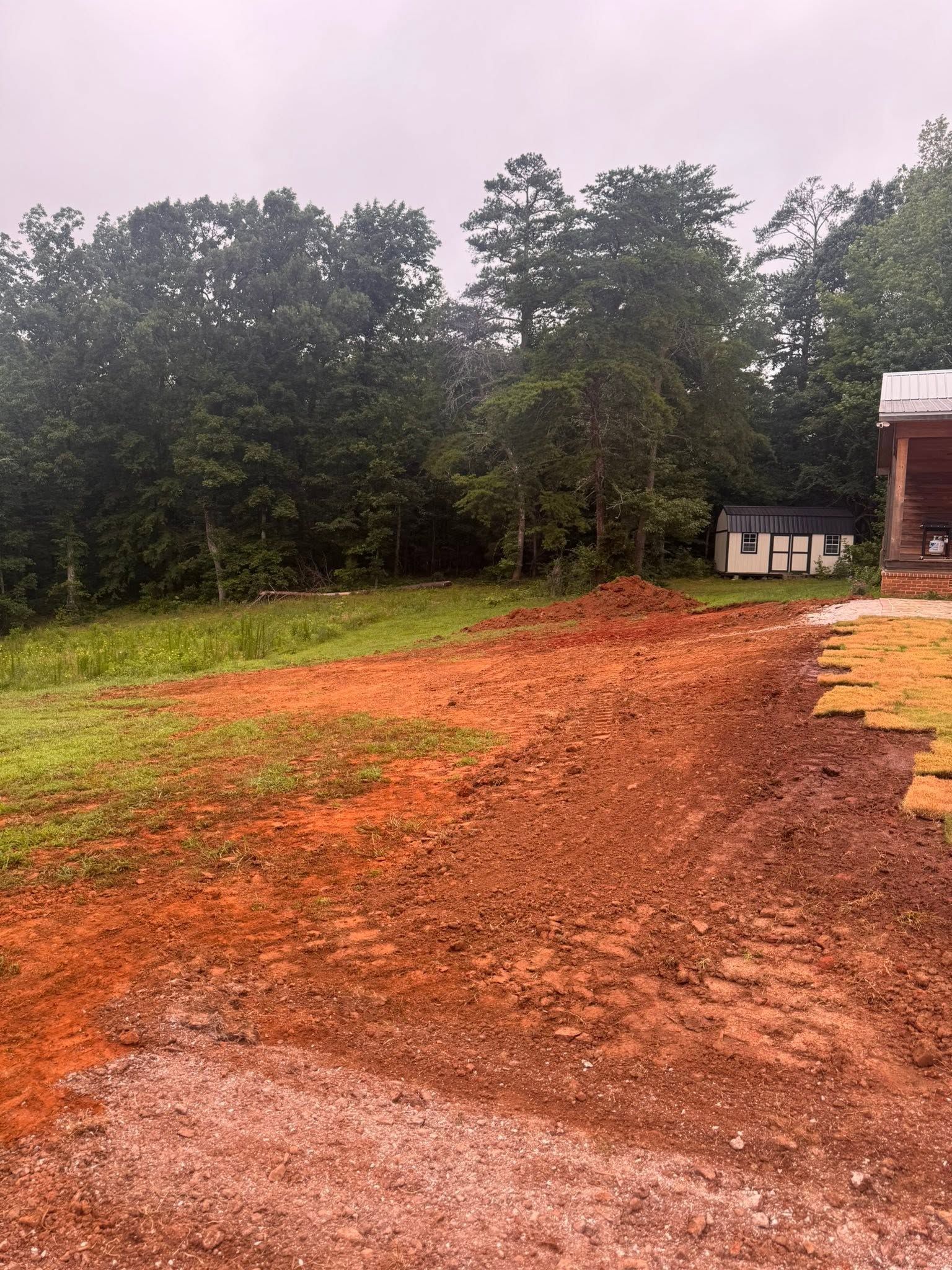 Muddy dirt lot beside trees and a small building under an overcast sky
