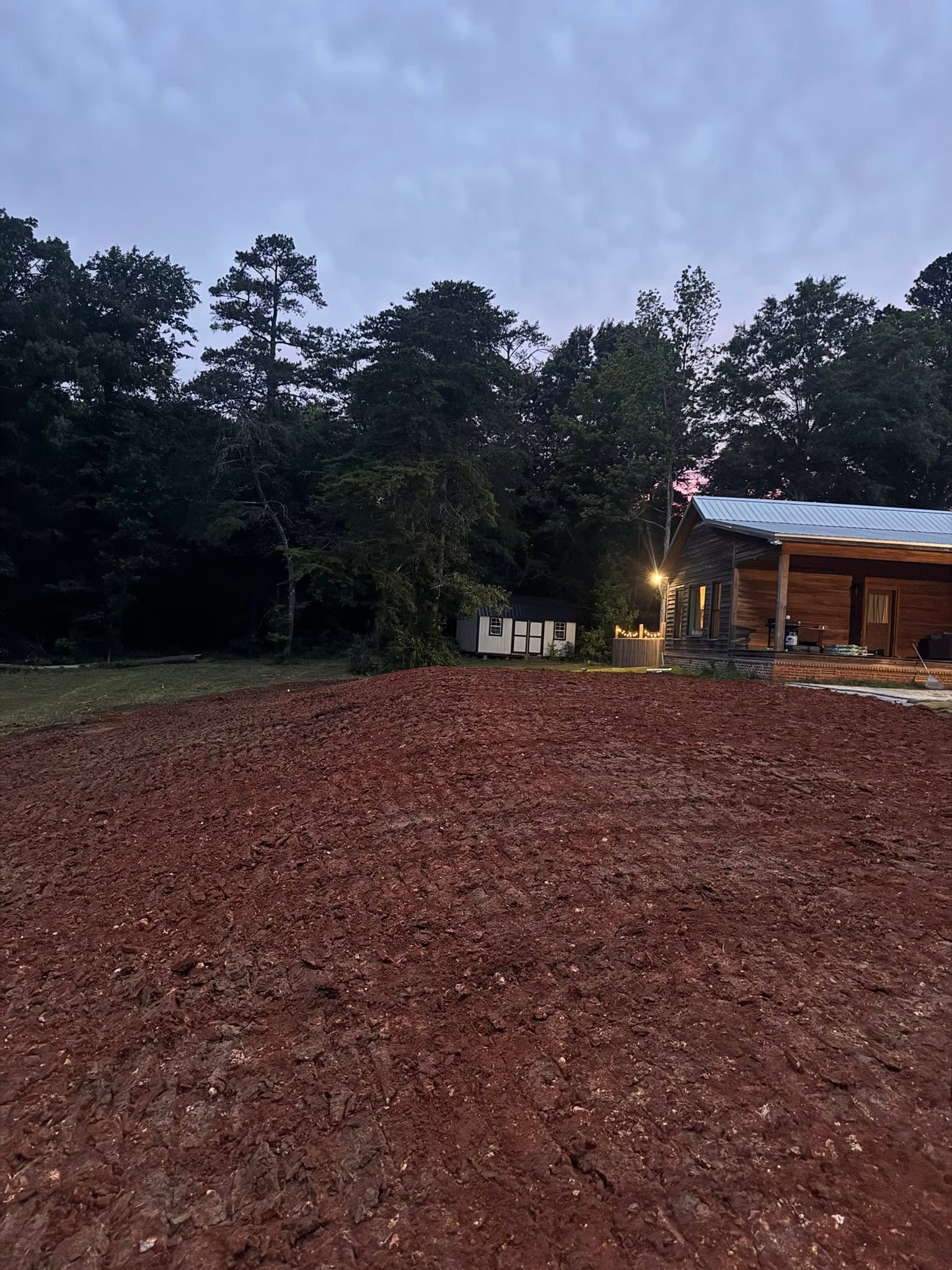 Wide red dirt clearing beside trees at dusk, with a small lit wooden cabin on the right.