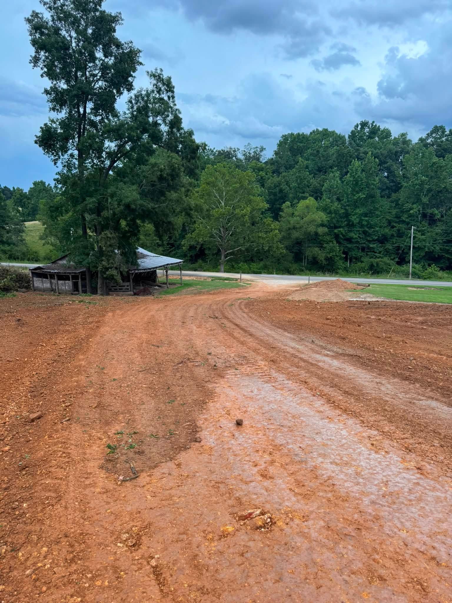 Dirt road curving through a wooded area under a cloudy sky, with a small building at left.