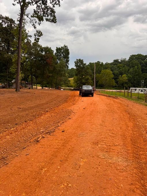 Car on an orange dirt road beside trees and a grassy field under a cloudy sky