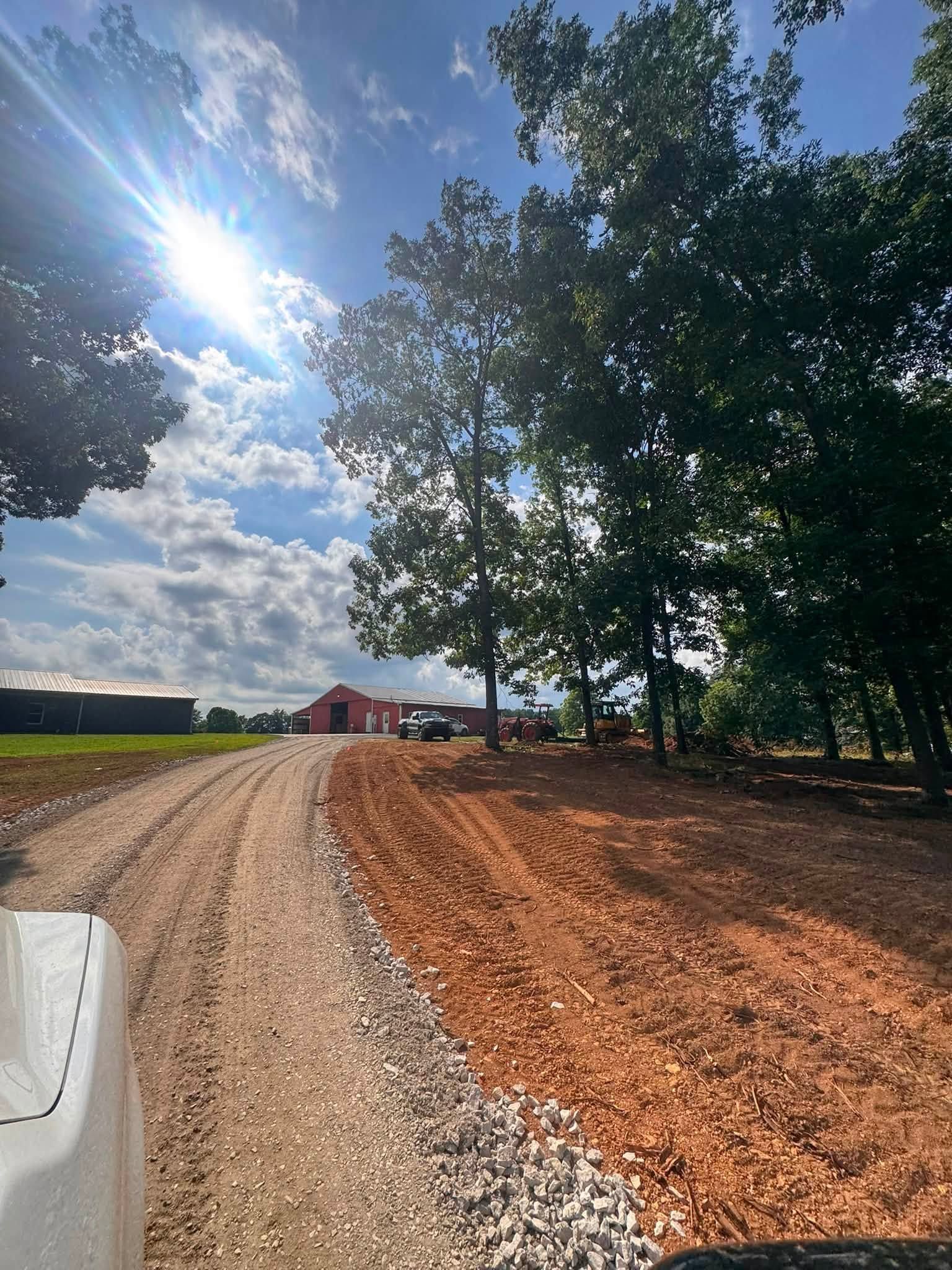 Sunny gravel driveway curving past trees toward a red barn under a blue sky with clouds