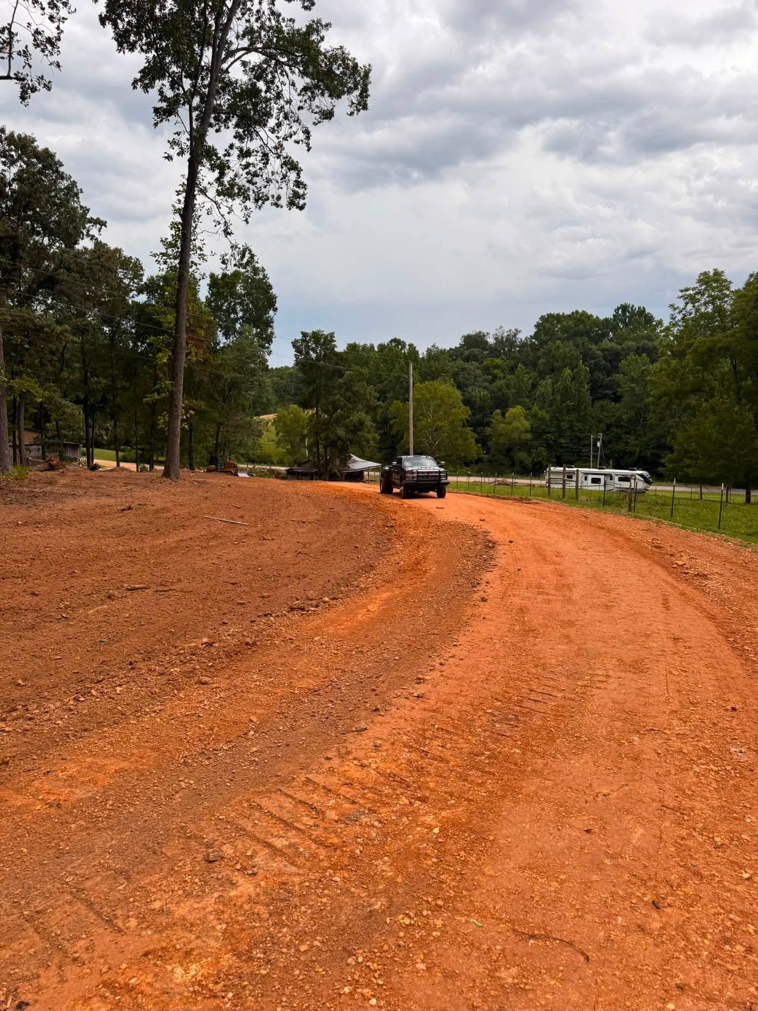 Curved orange dirt road through a wooded area under cloudy skies, with a parked car in the distance