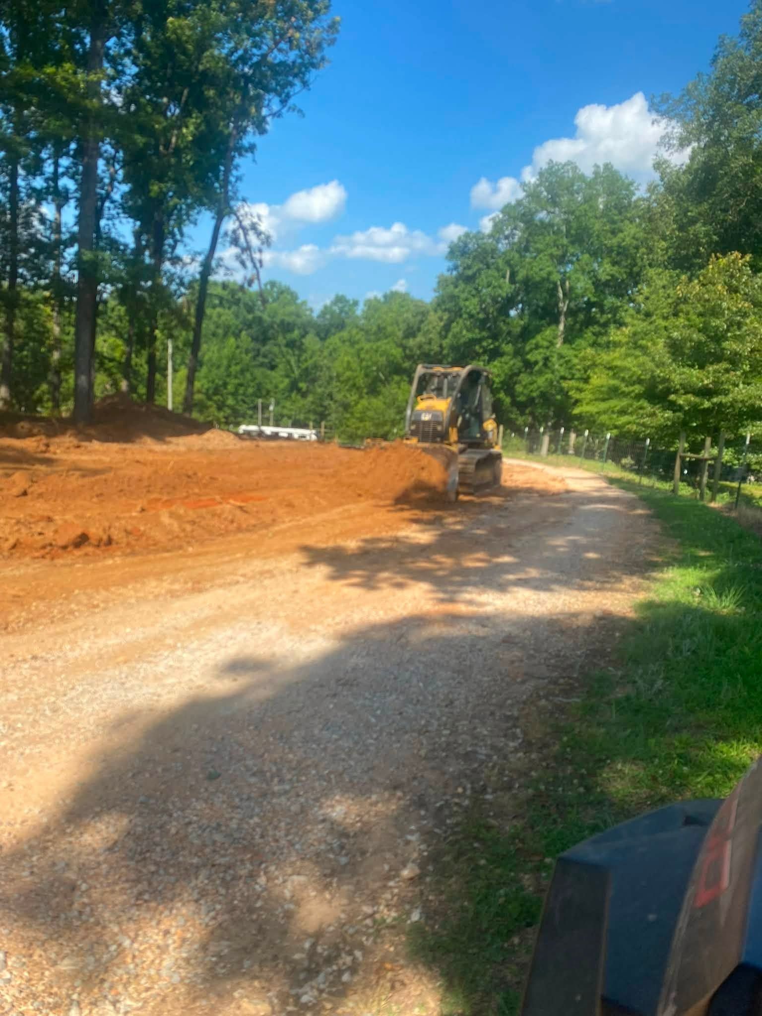 Bulldozer grading a dirt road beside trees under a bright blue sky