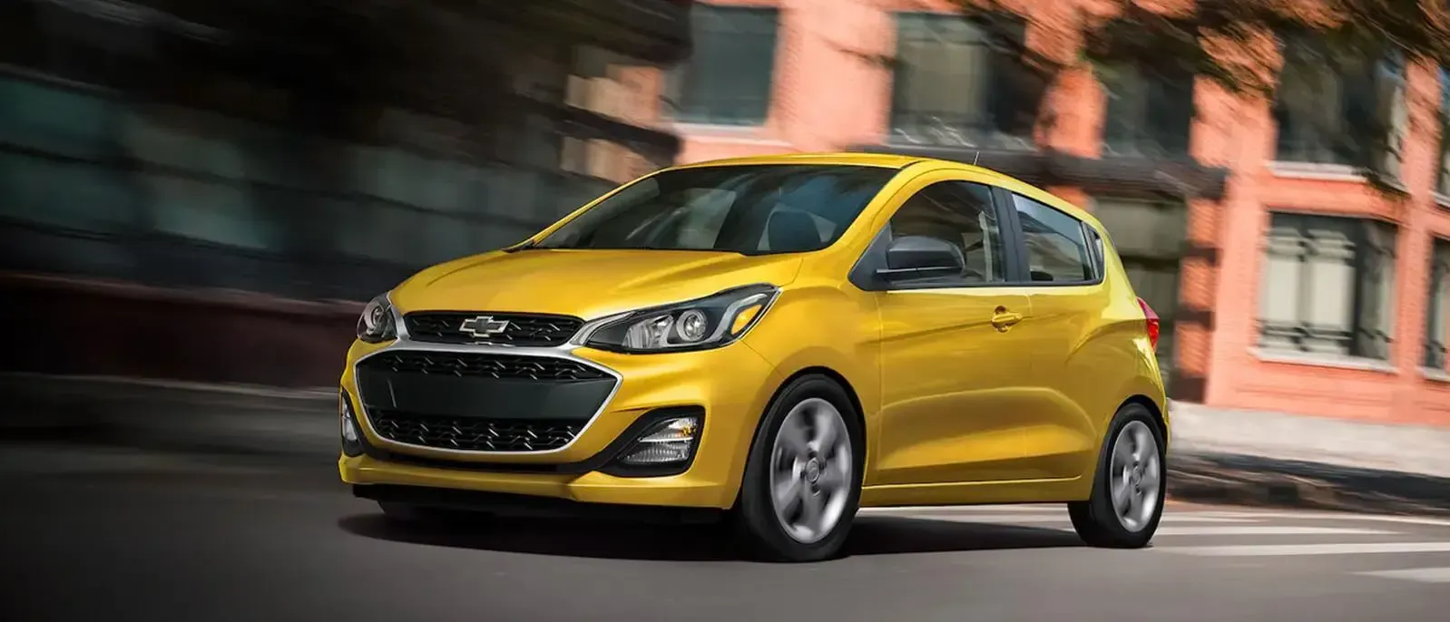 A yellow chevrolet spark is driving down a city street.