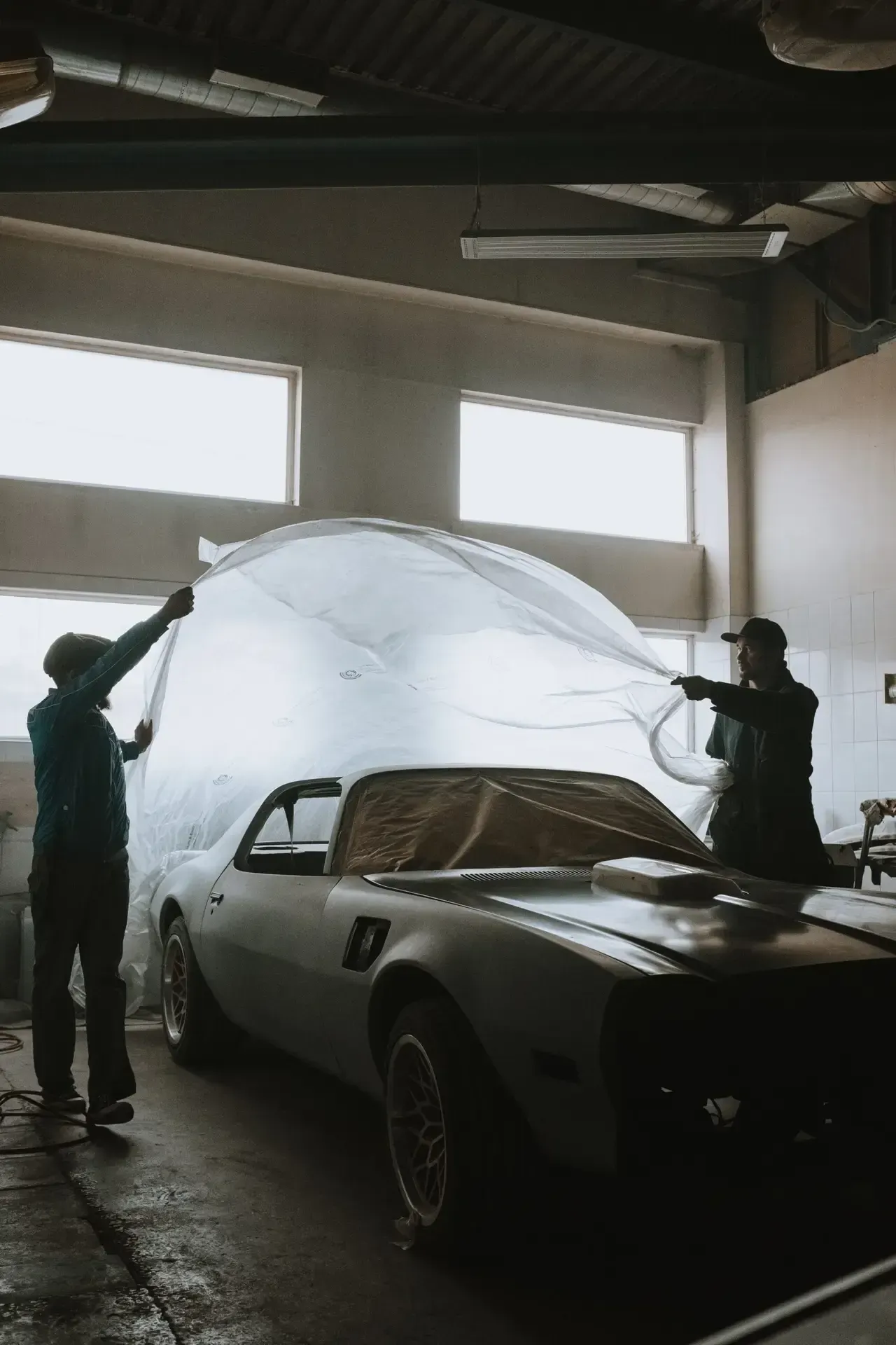 Two men are covering a car with a tarp in a garage.