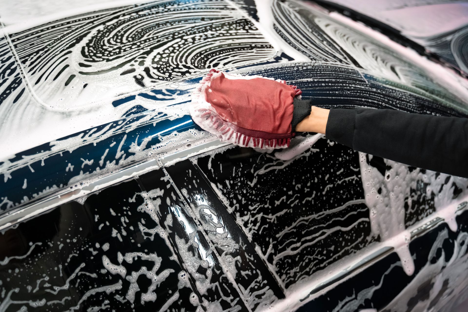 Gloved hand washing a black car with a red sponge, covered in soap suds