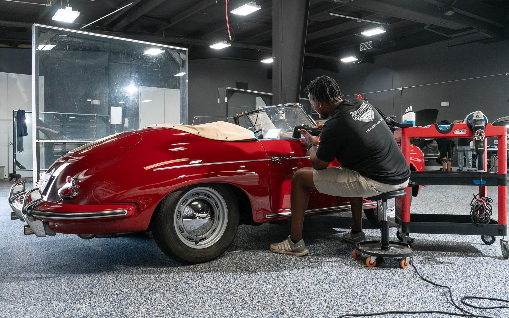 A man is sitting on a stool working on a red sports car in a garage.