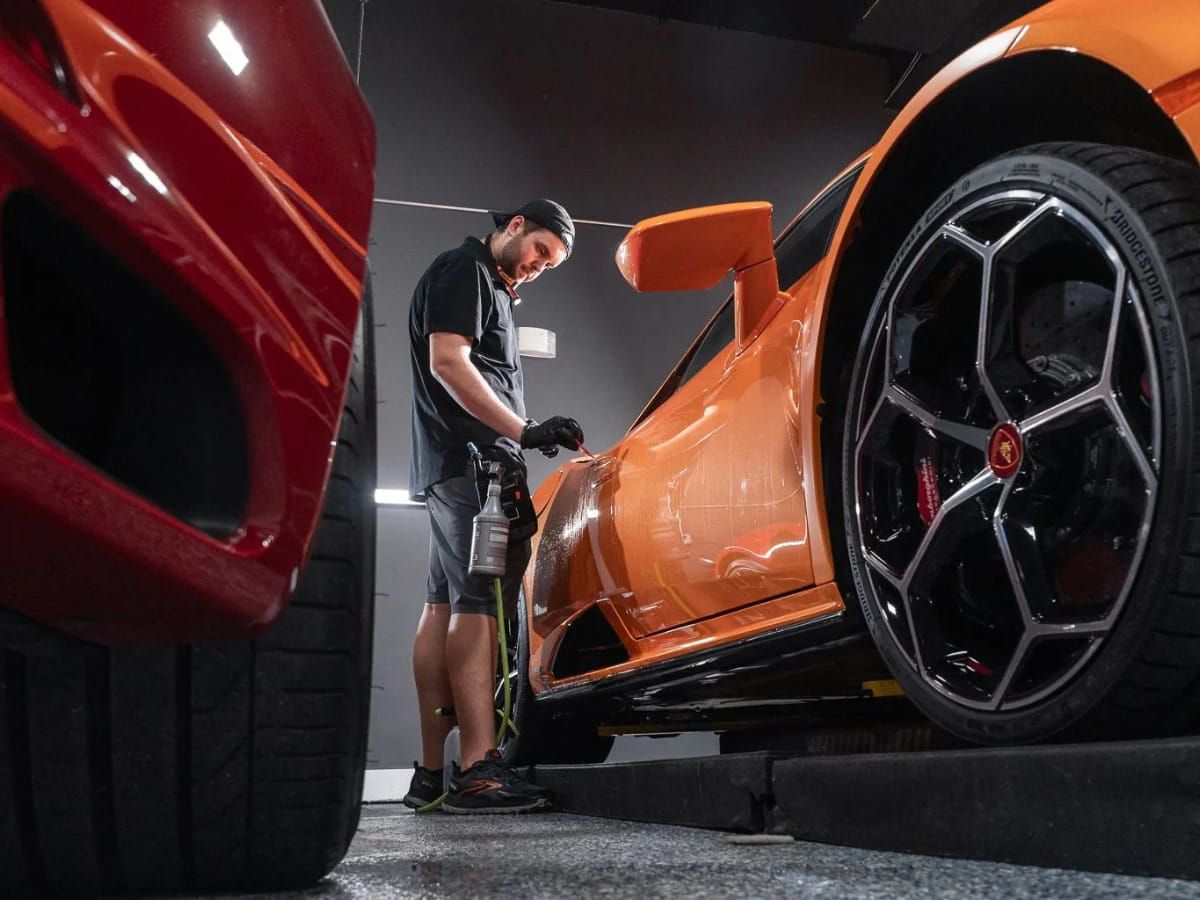 Technician working on the side of an orange sports car beside a black alloy wheel.