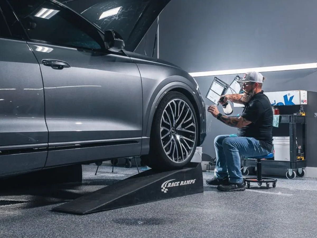 Technician seated beside a silver sedan while working near its front wheel inside a detailing shop.
