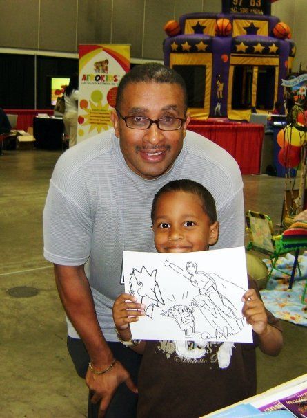 Man and young boy smiling, holding a Superman coloring page. They stand in front of a bounce house.