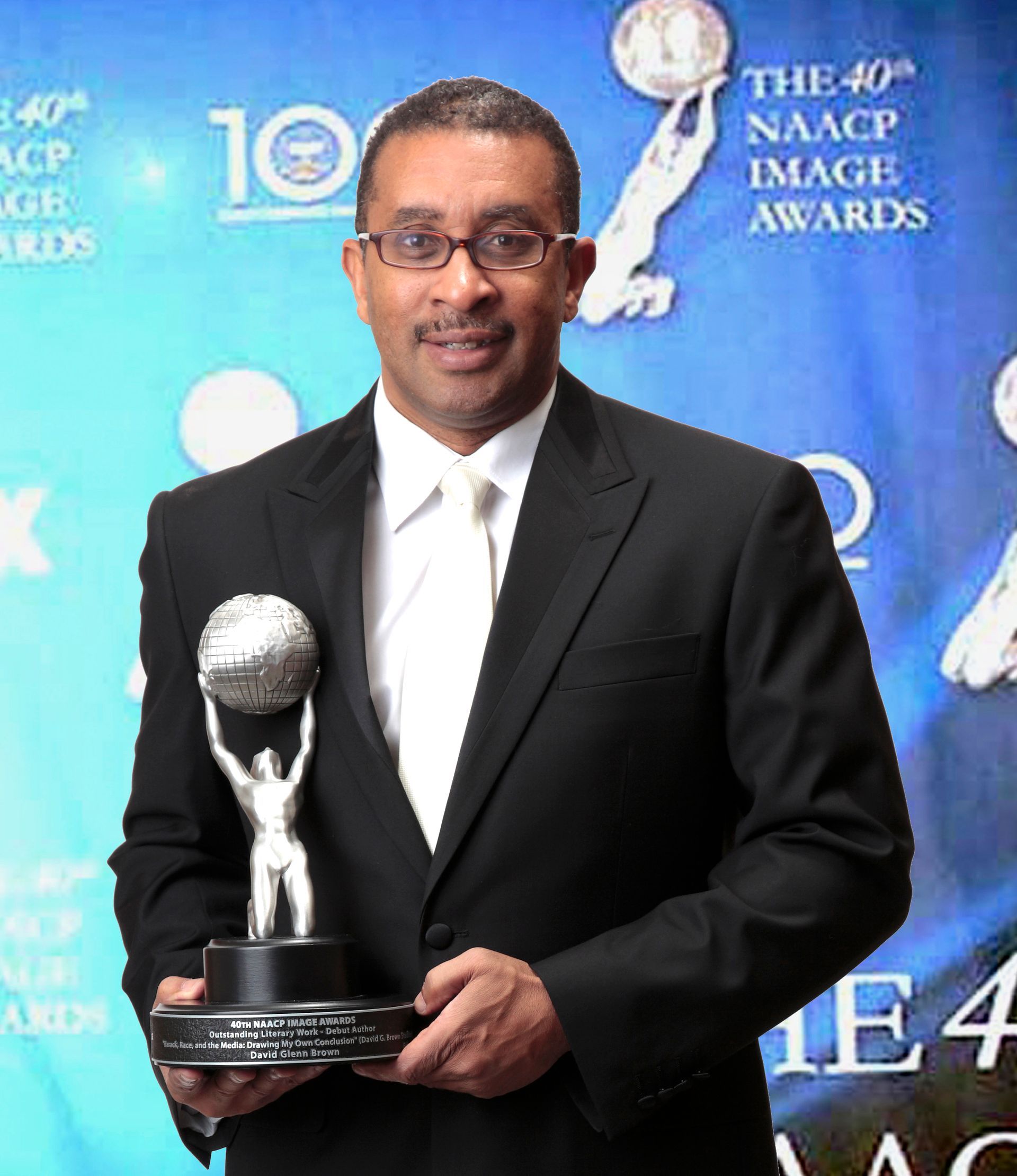 Man in a suit holds an NAACP Image Award. He wears glasses and smiles in front of a backdrop.