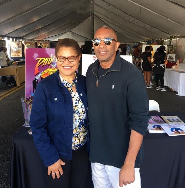 Two people smiling, posing together at an outdoor event. Woman in glasses and denim jacket; man in sunglasses and sweater.
