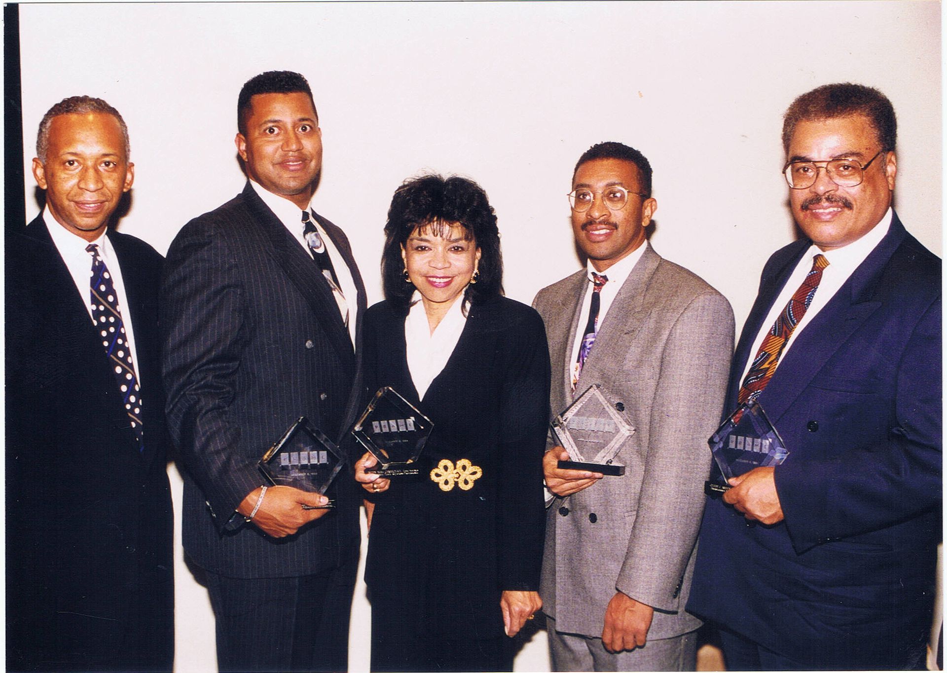 Five people in suits hold awards, smiling, standing in front of a white wall.